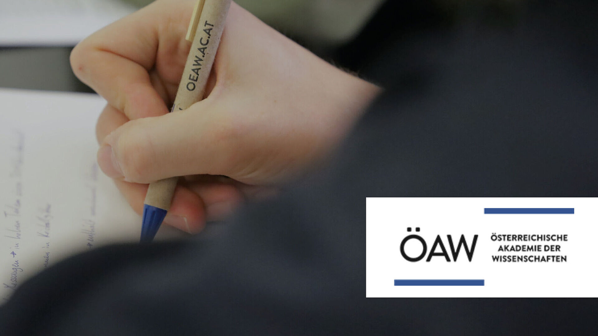 Kind Schreibt auf ein Blatt mit einem Kugelschreiber mit dem ÖAW Österreichische Akademie der Wissenschaften Logo